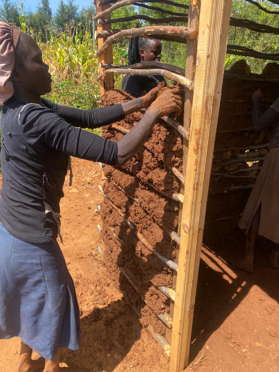 Here is the rewritten caption: Two individuals are constructing a small mud hut in a rural area. In this side view image, ...